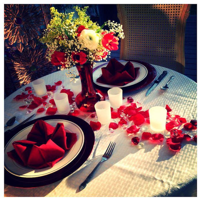 ideja romantičnega obroka z dvema rdečima prtičkoma na krožnikih za mizo v cvetnih lističih in svečah