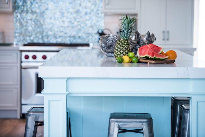 beyaz bir mutfakta vurgu olarak gök mavisi merkezi ada, gri bar tabureleri, tropikal meyveler, deniz kenarı dekoru