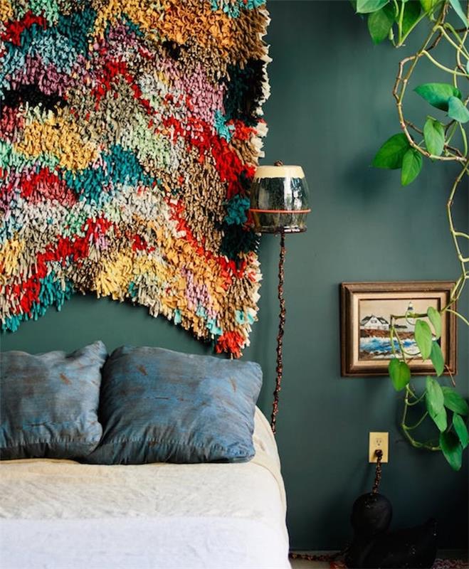 primer pisanega vzglavja iz tkanine, temno sivo zelena stena, bež in bela posteljnina ter modre okrasne blazine, zelena rastlina