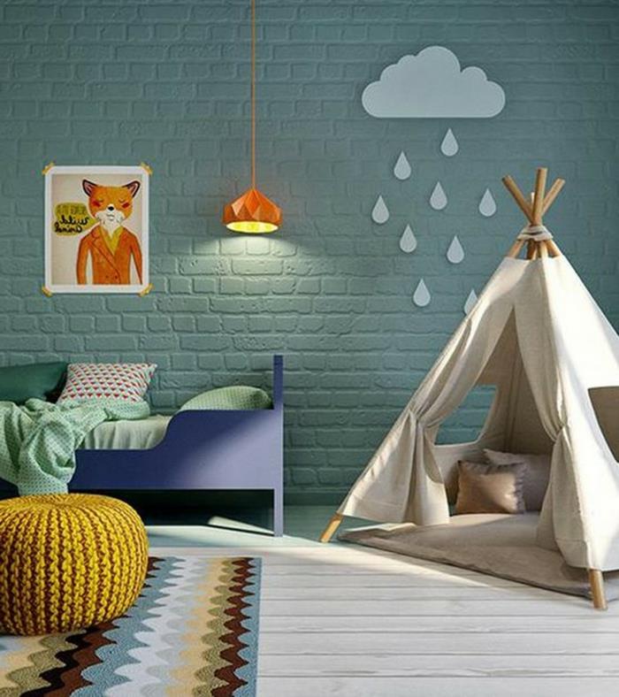 žalia plytų siena, vaikiškas triukas, geltona megzta taburetė, mėlyna vaikiška lova, įrėmintas piešinys ir oranžinė lempa