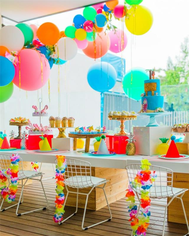 baltos kėdės, baltas stalas, pakabinami balionai, pyrago stovas su mėlynu pyragu ir auksiniai padėklai su keksiukais