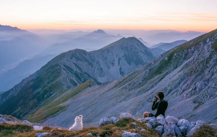 fotografas, sėdintis uolos pakraščiuose, kalnai su baltomis uolomis, dangus oranžinės ir mėlynos spalvos, baltas šuo