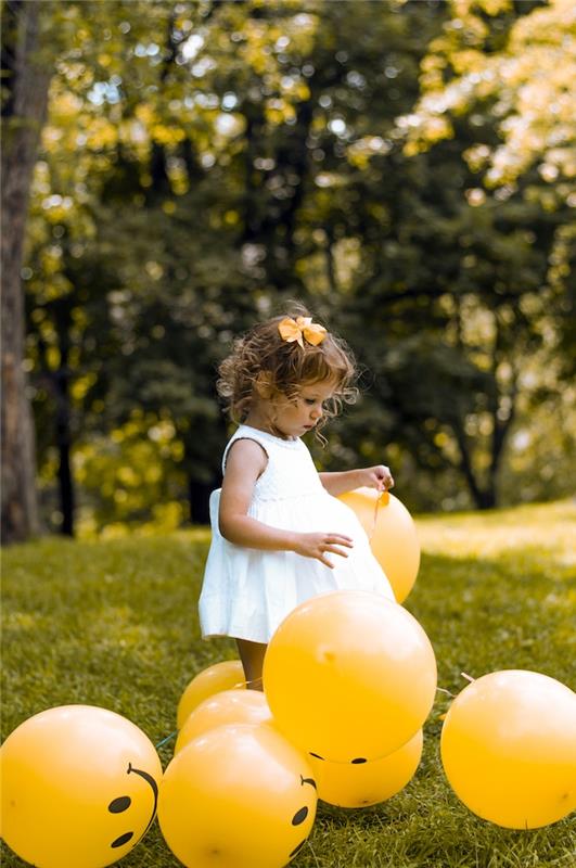 Pirmojo gimtadienio vakarėlis su geltonais balionais, sodo mergaitė, žaidžianti su balionais, maža mergaitė balta suknele, garbanotų plaukų šukuosena, mielas geltonas drugelis