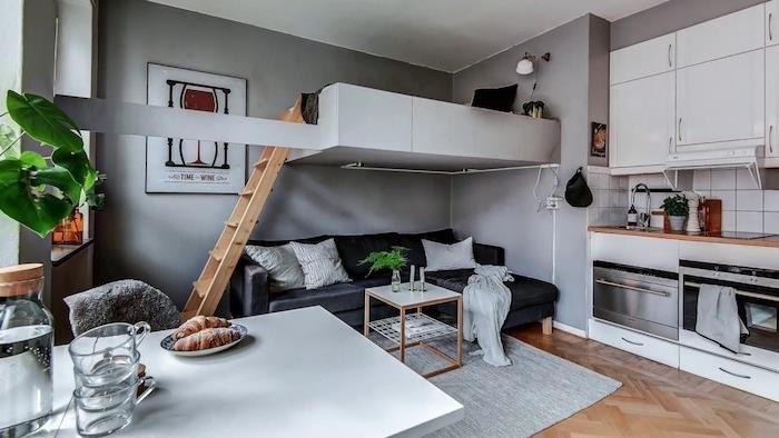 pilka sofa ir svetainės stalas po balta aukšta lova, pilkos sienos ir virtuvėlė baltai studijai, baltas valgomojo stalas