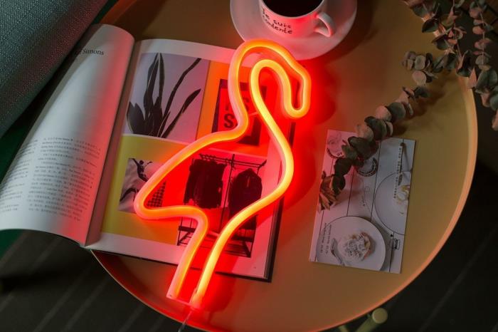 neonska svetilka v obliki svetlečega flaminga, majhna okrogla nočna omarica z modno revijo, razglednica in majhna bela skodelica za kavo, izviren okrasni predmet, rožnati okras flaminga
