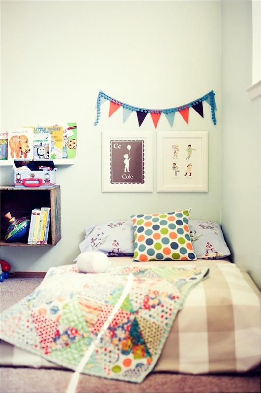 lova ant grindų, papuošta spalvinga antklode ir pagalvėlėmis, pilkas kilimas, baltos sienos, naktinis stalas iš perdirbto medžio dėžės, galvos apdangalas dekoratyviniuose rėmuose, minimalistinė vaikiška knygų spinta
