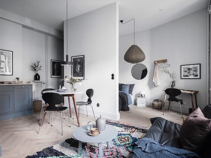 idėja, kaip įrengti erdvią studiją su siena atskirtu miegamuoju, svetainės erdve su juoda odine sofa, apvaliu žurnaliniu stalu, originaliu kilimu, stalu apsupta skandinaviškų kėdžių