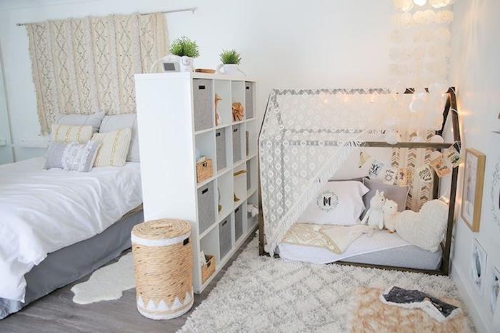 Įrengtas suaugusiųjų miegamasis su „Montessori“ erdve, atskirta lentyna, miegamoji lova ant grindų, papuošta žaislais ir pagalvėlėmis, lova su pilka ir balta lina