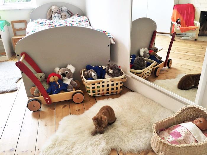 žema pilka lova su spalvinga antklode, sieniniu veidrodžiu, minkštu kilimu su katinu viršuje, šviesaus medžio parketas, žaislų krepšeliai