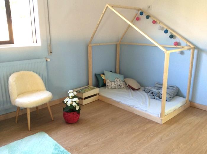 otroška kabina s svetlo leseno posteljo, spalnica montessori, dvobarvna stena v pastelno modri in beli barvi, venec barvitih pomponov na vrhu kabinske postelje