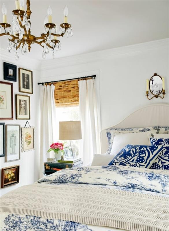 dekor podeželske spalnice, belo perilo z modrimi vzorci, stenska dekoracija s fotografijami in risbami, baročni lestenec, svetlo bele zavese, vintage nočna omarica