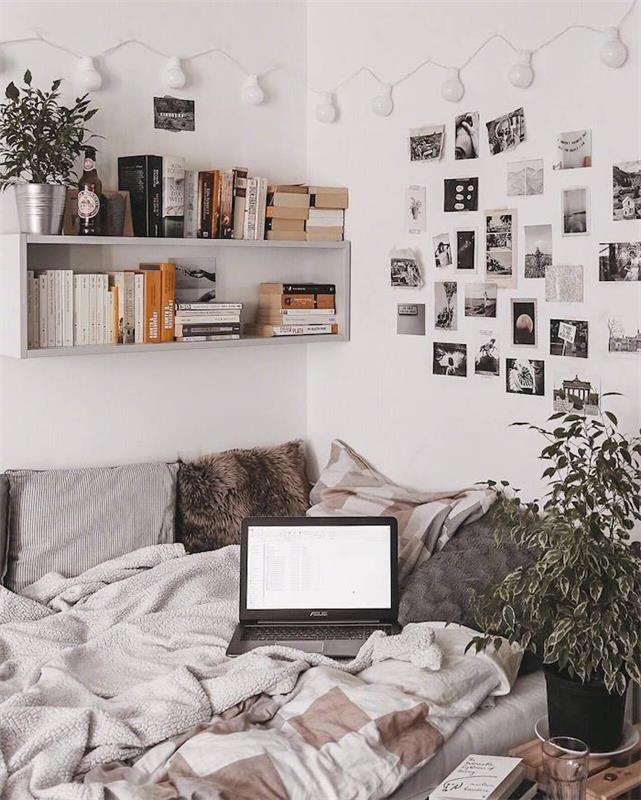 kampinė lova su rudomis ir pilkomis pagalvėmis ir lovų komplektais, balta sieninė lentyna su knygomis ir augalu, juodos ir baltos nuotraukų sienos, girliandos šviesa
