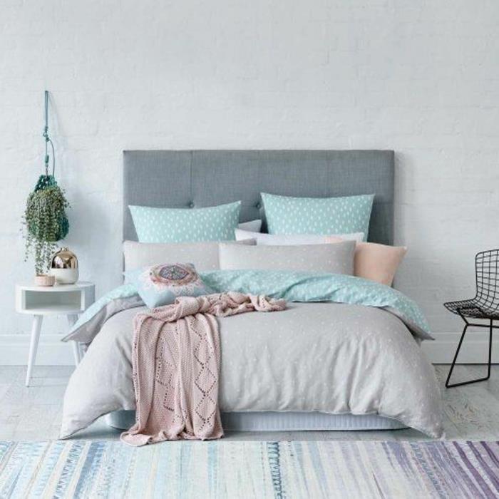 pilka lova, padengta pasteliniu vandens žaliu, pilkos ir rožinės spalvos patalyne, dėvėtos spalvos dryžuotas kilimas, baltų plytų siena, pilka galvūgalis, kabantis augalas