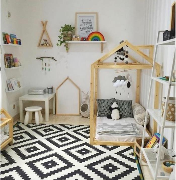 baltos ir juodos spalvos kilimas, geometriniai raštai, medinė lova su lova, baltas čiužinys, juodos ir baltos pagalvėlės, baltas stalas ir taburetė, knygų sieninė lentyna, mobilusis kūdikis, Montessori miegamasis