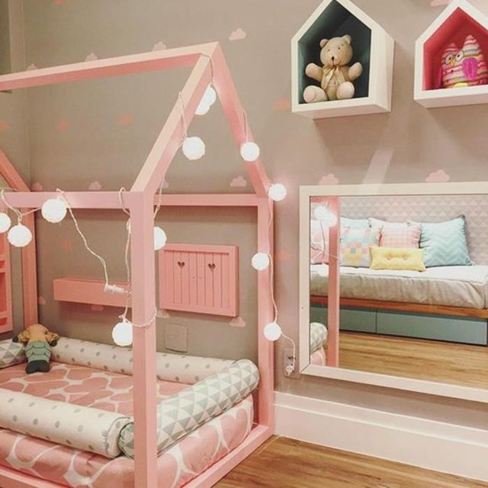rausva rožinė lova, šviesi girlianda, rožinis čiužinys, žemas veidrodis, žaislų saugykla, mažas medinis žaidimų namelis, šviesus parketas, Montessori pedagogika, miegamojo išdėstymas