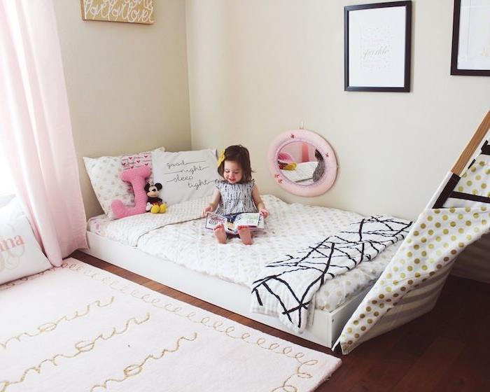 „Montessori“ lova ant grindų su nameliu „teepee“ ant rudo parketo, baltas kilimas su tekstu, smėlio spalvos sienos, merginos kambario išdėstymas