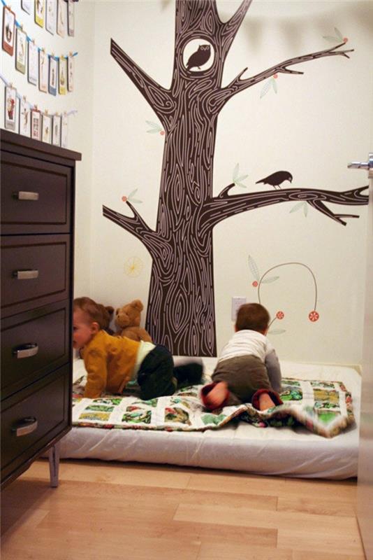 stene otroške spalnice v kremno beli barvi, veliko rjavo drevo, pobarvano s sovo in krokarjem na vejah, otroška posteljica brez palic