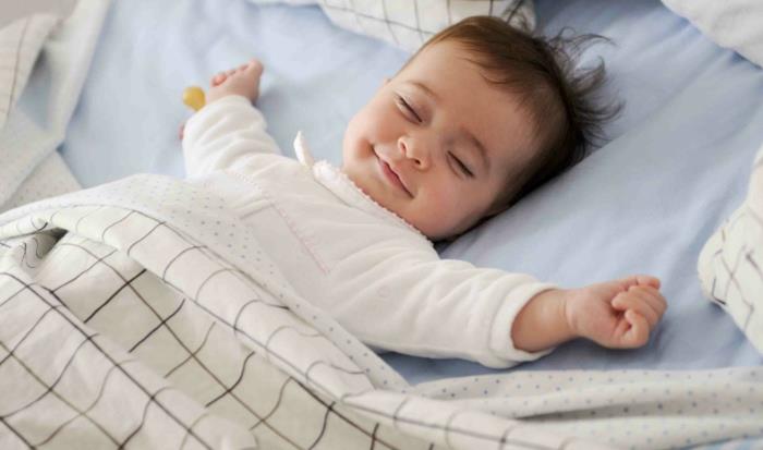 dojenček, ki srečno spi v postelji, jaslice brez palic, pastelno modro -belo perilo s ploščicami iz taupe, malček se nasmehne v postelji