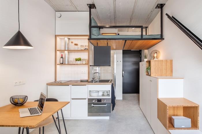 lesena in bela studijska kuhinjska niša z mansardno posteljo s sivo žimnico in blazino, betonska tla, lesena in kovinska miza
