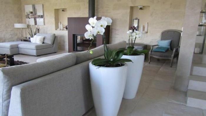 didelės keraminės vazos baltos spalvos, su pasodintomis baltomis orchidėjomis, šalia šviesiai pilkos sofos, svetainės spalvų idėjos, dėmėtos šviesiai smėlio spalvos sienos, tamsiai rudas židinys