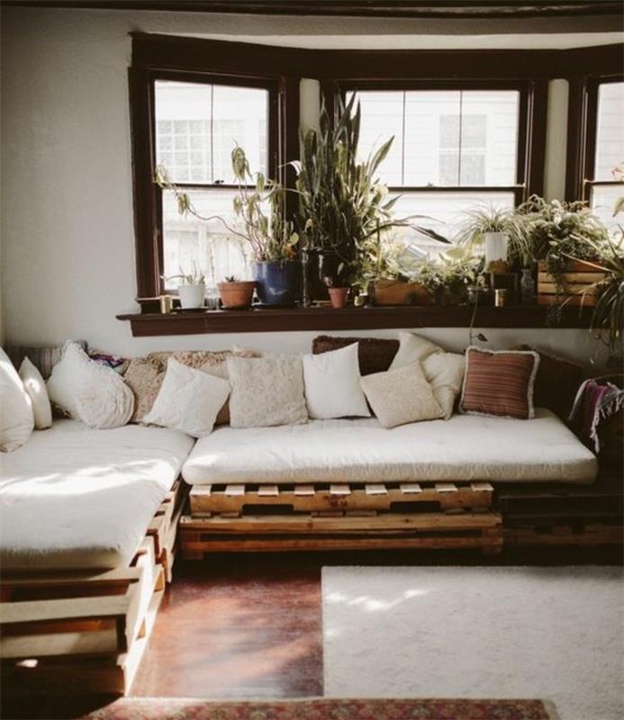 padėklų sofa, padengta baltomis, smėlio ir rudomis pagalvėmis, kambario su rudomis grindimis, šviesiai smėlio kilimėliu ir palange su daugybe vazoninių augalų viduje