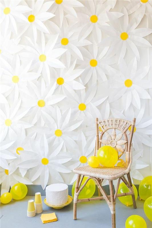 bir parti için bir duvar süsleme fikri, beyaz ve sarı kartondan büyük papatyalar, kolçaksız dokuma hasır sandalye, büyük bir çiçek deseniyle süslenmiş sırtlık, birkaç sarı balon, gri çinilerle kaplı zemin