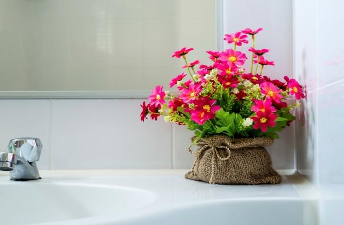 vonios kambario augalas be šviesos, gėlės fuksijos spalvos, puodelis, apsirengęs siuvinėtu drobės maišeliu, kambarinių augalų atspalvis, baltos kriauklės kampas, papuoštas gėlėmis