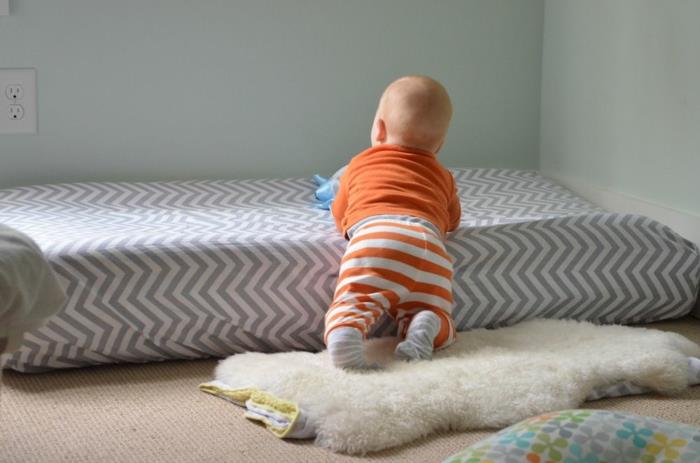 montessori odası, beyaz ve taupe çapraz çizgili yatak, reseda duvarlar, bej halı, parmaklıksız yatağına tırmanan bebek