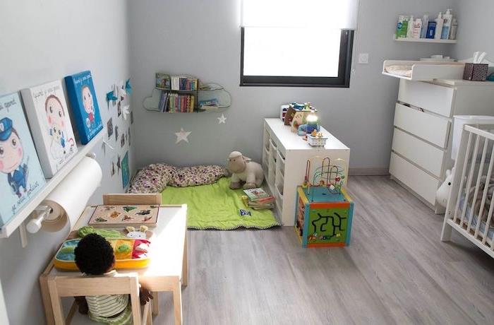 Montessori erdvės idėja tradiciniame miegamajame su žaliu žaidimų kilimėliu, žaislais ir knygų lentyna, mediniu stalu ir žemomis kėdėmis