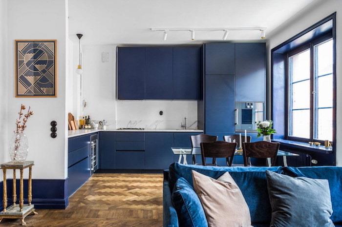 polnočno modra kuhinja, odprta za dnevno sobo s temno modrim kavčem, lesenimi tlemi, belimi stenami in temno modro visoko in nizko omarico v barvni karti Pantone