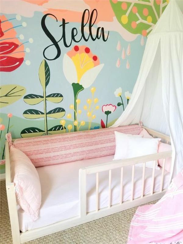 orman, bitki, çiçek ve güneş desenleri ile pastel renklerde çizilmiş duvarlar, küçük kız Stella'nın yazıt adı, kısmi çubuklu Montessori yatağı, bej halı, beyaz ve pembe kumaşta cibinlik