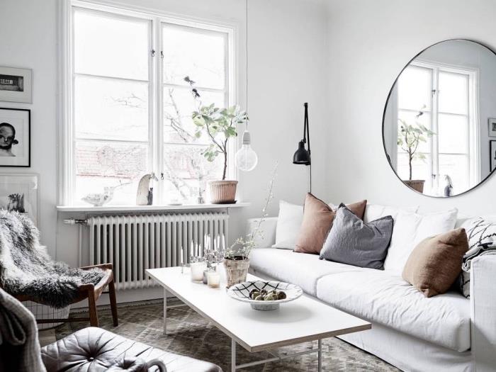 Modern bir oturma odasında İskandinav mobilyaları, beyaz kanepe, gri ve kahverengi minderler, beyaz sehpa, geometrik şekilli gri halı, gri hayvan derisi sandalye, beyaz iç mekan, yuvarlak ayna