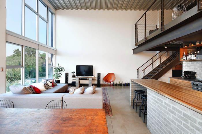 industrijsko leseno-betonsko blok TV pohištvo, tla iz keramičnih ploščic, sivi kotni kavč, orientalska preproga, odpiranje v kuhinjo, medetaža