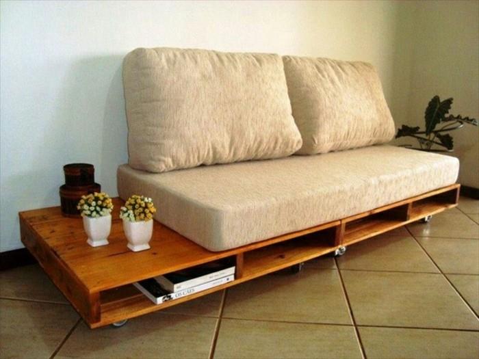 retro stiliaus „pasidaryk pats“ sofa, pagaminta iš šlifuotų medinių padėklų, šiltai rudos spalvos, su ratukais ir laikymo vietomis, smėlio spalvos sofos pagalvėlėmis ir vazoniniais augalais
