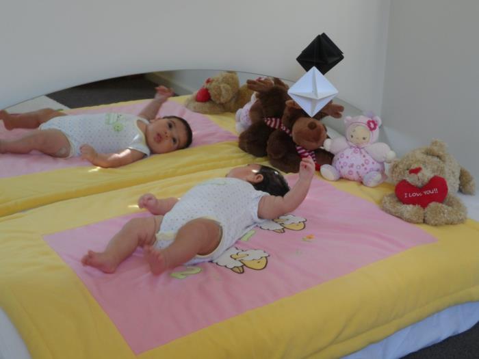 dojenček leži na posteljici brez palic, rumena in roza odeja, polnjene živali, pisani kartonski predmeti, ki visijo nad dojenčkom