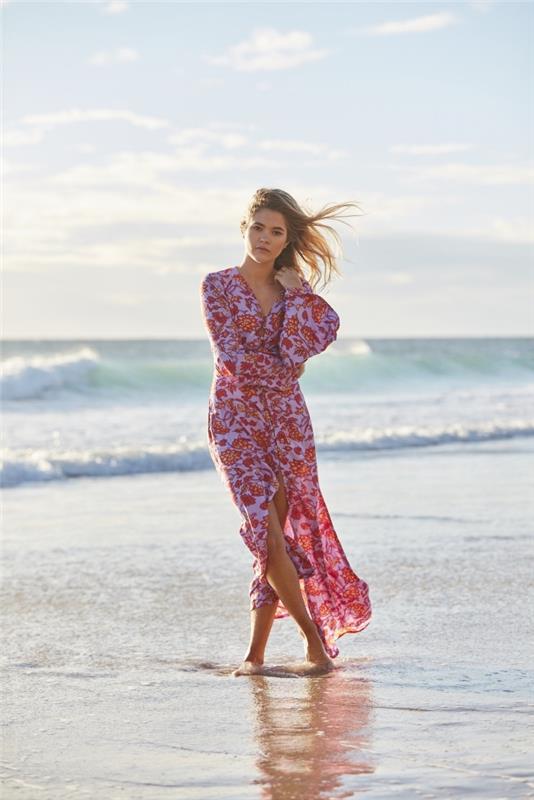 jauna moteris paplūdimyje, apsirengusi šviesia vasarine suknele raudonomis ir rožinėmis spalvomis asimetriško dizaino ilgomis rankovėmis