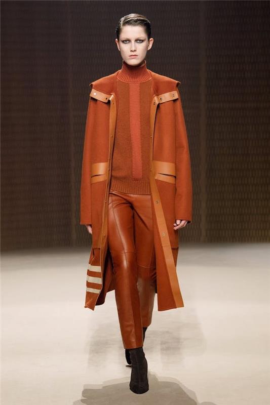 trendi oblačil, manekenka, ki hodi po vzletno -pristajalni stezi, oblečena v rjave usnjene hlače, oranžni pulover in dolg plašč