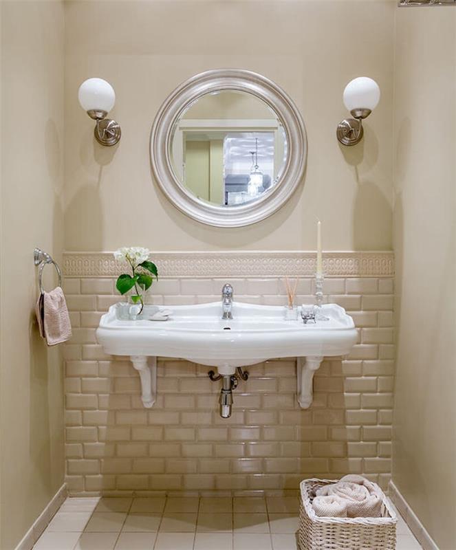 küçük bej bir banyoda eski beyaz lavabo, yuvarlak ayna, saklama sepeti ve duvar lambaları
