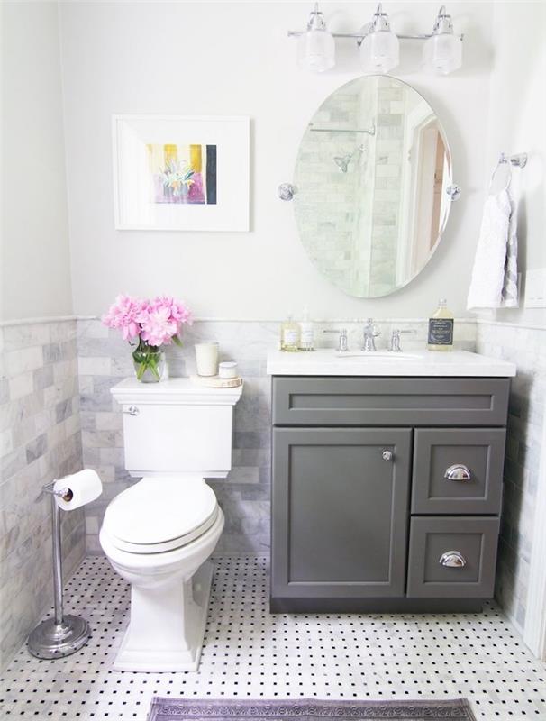 beyaz tuvalet, gri makyaj ünitesi, siyah noktalı beyaz yer karoları, oval ayna, banyo aydınlatması ile küçük tasarım banyo