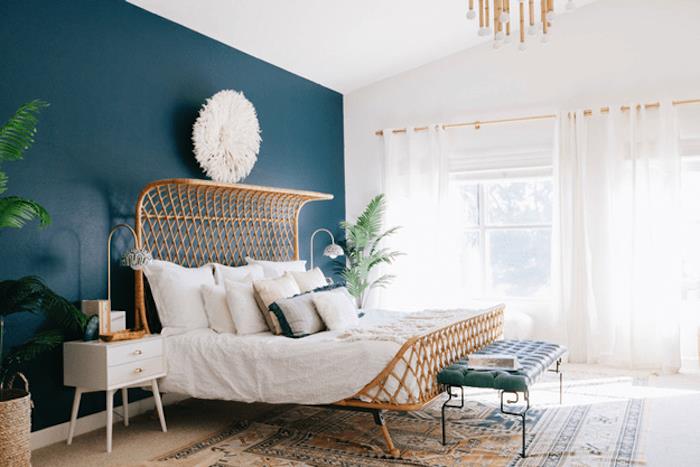 idėja, kaip įrengti miegamąjį, rytietišką stilių, medinę lovą, baltą patalynę, rytietišką kilimą, žalią augalą, naftos mėlynus dažus vienoje sienos dalyje, didelius langus ir baltas užuolaidas