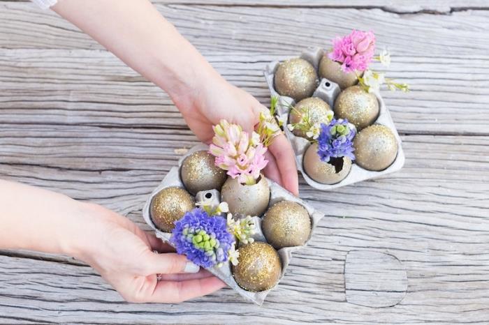 Velykų stalo dekoravimo idėja su tuščiais kiaušiniais ir nudažyta aukso spalvos blizgučiais vazų su gėlėmis, kiaušinių dėžutės su gėlėmis ir dekoruotais lukštais pavidalu