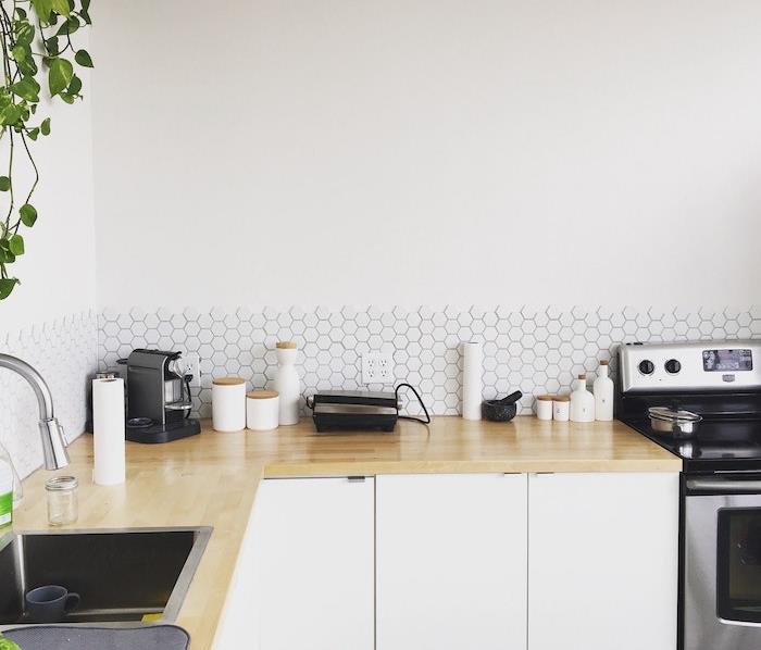 mažas baltos spalvos virtuvės modelis su šviesaus medžio stalviršiu, baltos spalvos plytelėmis, nerūdijančio plieno prietaisai