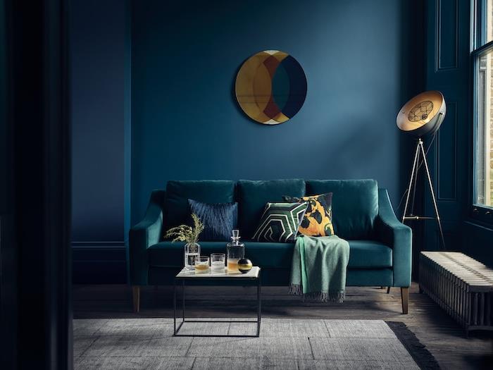 benzino mėlynos svetainės idėja su povo mėlyno atspalvio sofa, žalia antklodė, juodas minimalistinis stalas su baltu viršumi, tamsiai mėlyni sienų dažai, pilkas kilimas