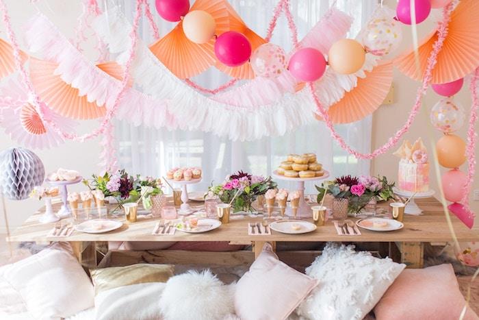 prašmatni bohemiška gimtadienio puošmena su baliono puošmena, vėduoklės girlianda, kaimiškas prašmatnus stalas, papuoštas gėlėmis, ledų spurgos ir pyragai, bohemiškos pagalvėlės ant grindų