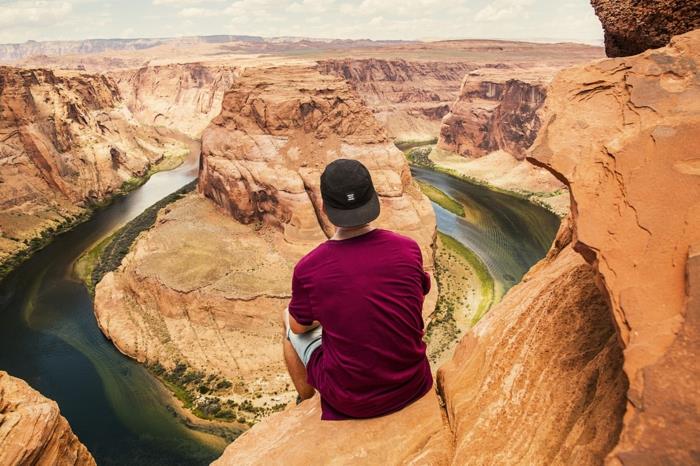 rojaus kraštovaizdis, vyras su bordo spalvos marškinėliais, sėdintis ant uolos krašto virš kanjono, vanduo su žaliais ir mėlynais atspalviais
