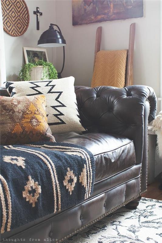 Actekų raštas, ruda odinė sofa, etninės kilmės kilimėlis ir pagalvėlės, šviesiai pilka siena