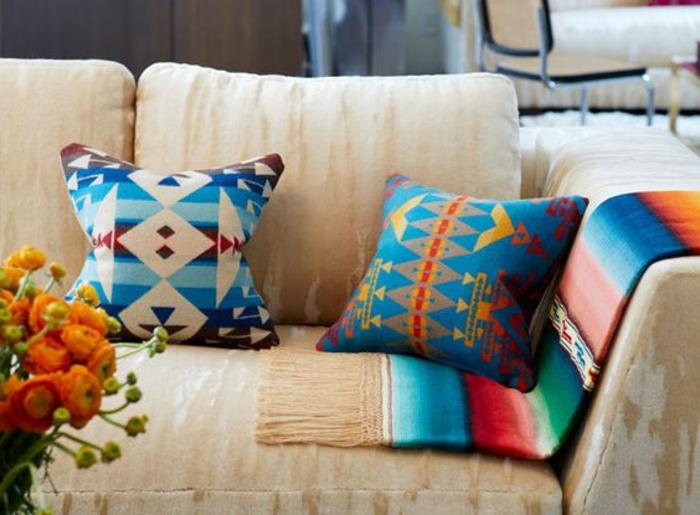 Actekų raštas ant pagalvėlių, pagalvėlės mėlynos ir oranžinės spalvos, smėlio spalvos sofa, oranžinių gėlių puokštė