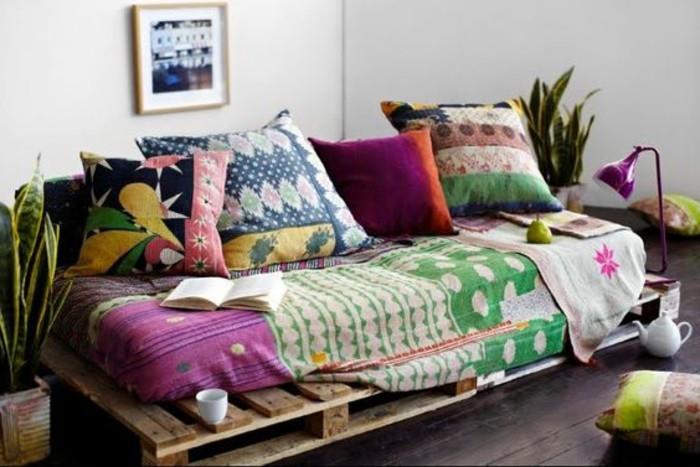 boho stiliaus padėklų sofa, padengta įvairiomis tekstilės medžiagomis ir pagalvėlėmis, įvairių spalvų ir raštų, du vazoniniai augalai netoliese