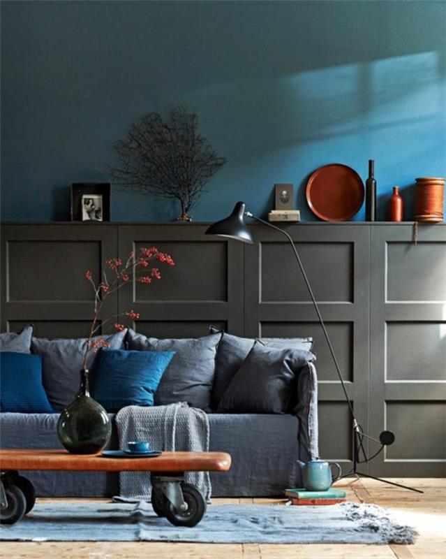 anties mėlyna siena, pilka sofa su pagalvėlėmis, pramoninis stalas, toršeras