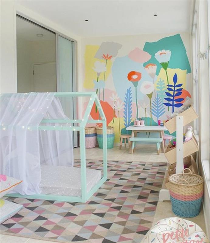 įvairiaspalvis trikampis kilimas, žalias Montessori žaidimų namas, baldakimas, gėlių rašto akcentinė siena, žaislų krepšelis, kavos staliukas, medinis suoliukas, taburetė, balta siena
